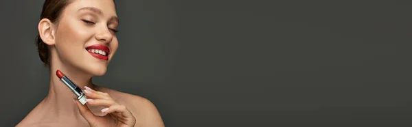 Allegra giovane donna con labbra rosse che tengono il rossetto e sorridono su sfondo grigio, banner per il trucco — Foto stock