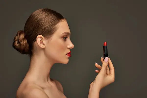 Vista lateral da bela jovem segurando batom vermelho no fundo cinza, tendências de beleza — Fotografia de Stock