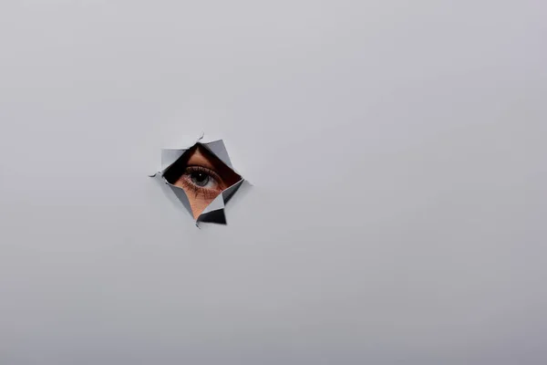 Tiro parcial de jovem mulher com olho azul olhando para a câmera através de buraco em fundo cinza rasgado — Fotografia de Stock