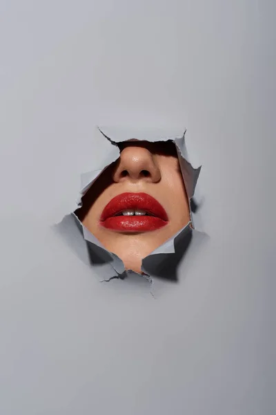 Cropped view of young woman with red lips near ripped grey background with hole, peeking — Stock Photo