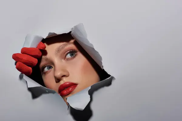 Attractive young woman with blue eyes and red lipstick peeking through hole in ripped grey paper — Stock Photo