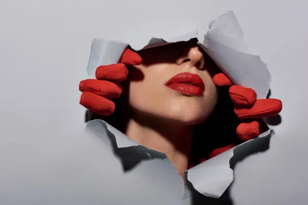 Cropped view of young woman with red lips in gloves ripping grey background and making a hole — Stock Photo