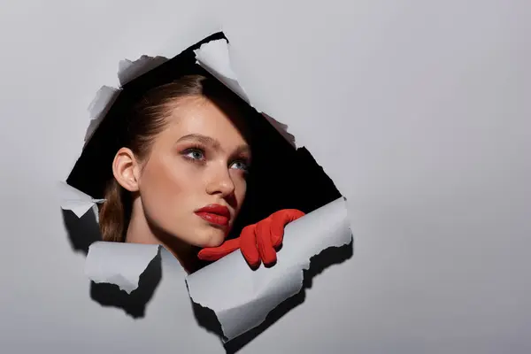 Pretty young woman with blue eyes and red lipstick peeking through hole in ripped grey paper — Stock Photo