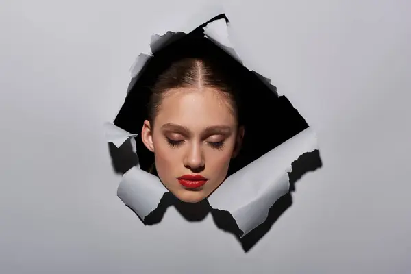 Pretty young woman with blue eyes and red lipstick peeking through hole in ripped grey paper — Stock Photo