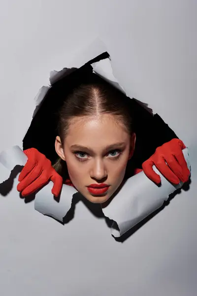 Jovem com batom vermelho e luvas espreitando através do buraco em papel cinza rasgado, maquiagem ousada — Fotografia de Stock