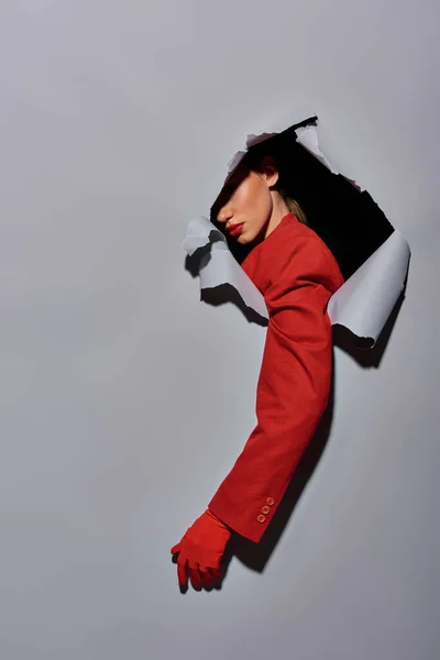Recortado tiro de mujer con manga roja y guante rompiendo a través de agujero en fondo gris, conceptual - foto de stock