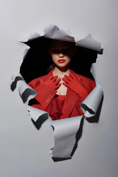 Привлекательная молодая женщина с смелым макияжем в красных перчатках позирует возле отверстия на сером фоне — стоковое фото