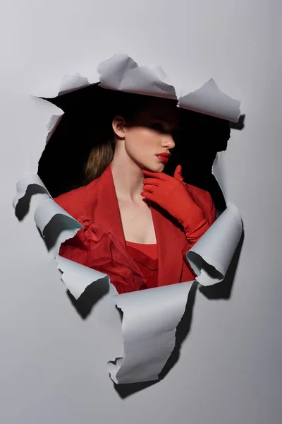 Glamourosa jovem com maquiagem ousada posando em luvas vermelhas perto de buraco no fundo cinza rasgado — Fotografia de Stock