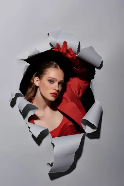 Atractiva joven con labios rojos y guantes rompiendo a través del agujero en el fondo gris, conceptual - foto de stock