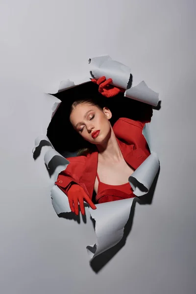 Bonita mujer joven con labios rojos y guantes rompiendo a través de agujero en fondo gris, conceptual - foto de stock