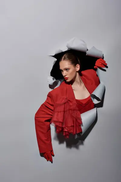 Bonita mujer joven en chaqueta roja y guantes rompiendo a través del agujero en el fondo gris, conceptual - foto de stock