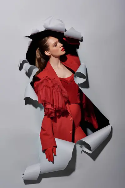 Femme élégante en blazer rouge et gants rompant trou dans fond gris déchiré, conceptuel — Photo de stock