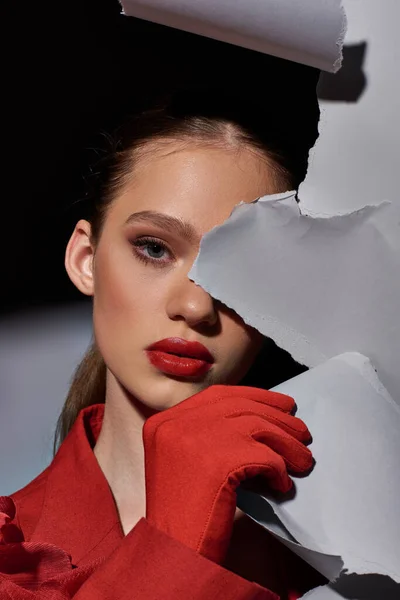 Attraktive junge Frau mit leuchtend roten Lippen posiert neben zerrissenem grauen Papier und blickt in die Kamera — Stockfoto