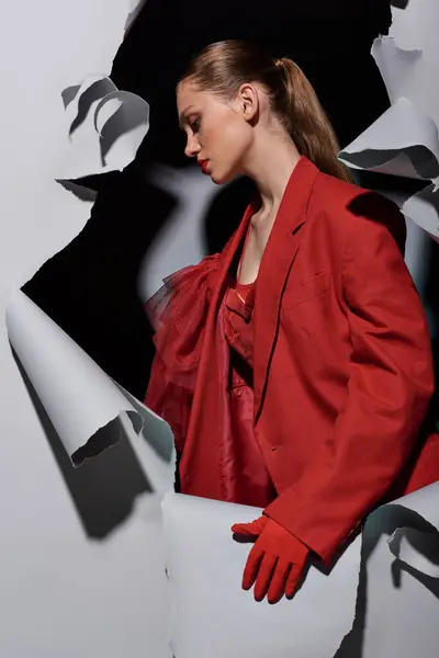 Vue latérale de la jeune femme à la mode en tenue rouge avec un maquillage audacieux debout près de fond gris déchiré — Photo de stock
