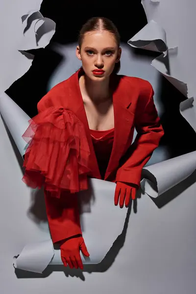 Mujer joven de moda en traje rojo con maquillaje audaz rompiendo a través de fondo gris con agujero - foto de stock