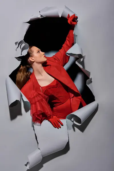 Jovem elegante em traje vermelho com maquiagem ousada rompendo fundo cinza com buraco — Fotografia de Stock