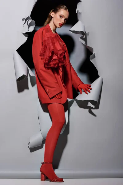 Mujer joven con estilo en traje rojo con maquillaje audaz rompiendo a través de fondo gris con agujero - foto de stock