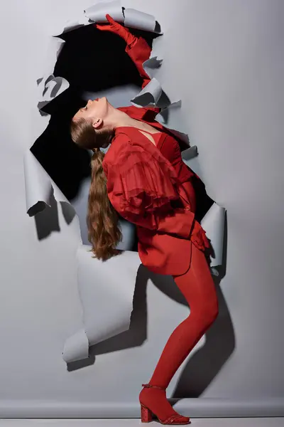 Mujer joven de moda en traje rojo con maquillaje audaz posando cerca de fondo gris roto con agujero - foto de stock