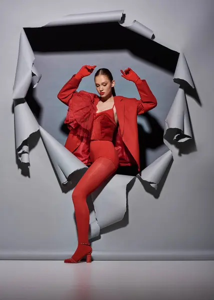 Могущественная молодая женщина в красной одежде с перчатками, прорывающимися сквозь порванный серый фон с отверстием — стоковое фото