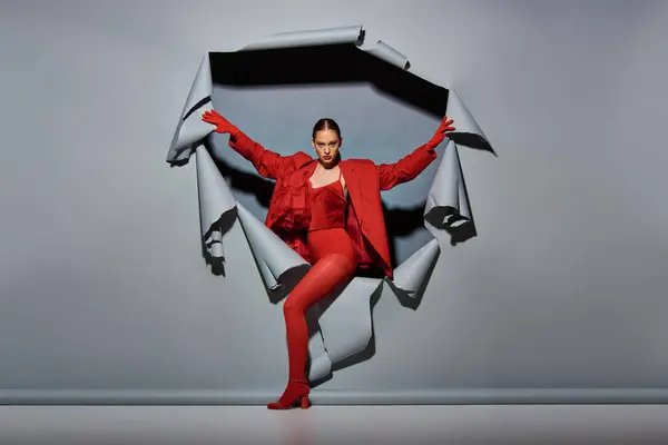 Selbstbewusste junge Frau im roten Outfit mit Handschuhen durchbricht zerrissenen grauen Hintergrund mit Loch — Stockfoto