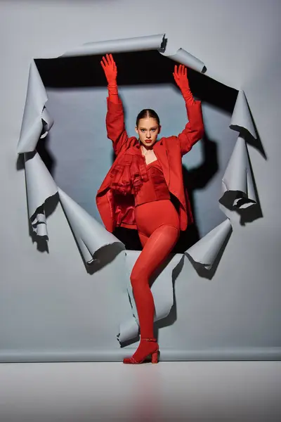 Уверенная в себе молодая женщина в красной одежде с перчатками, прорывающимися сквозь порванный серый фон с отверстием — стоковое фото