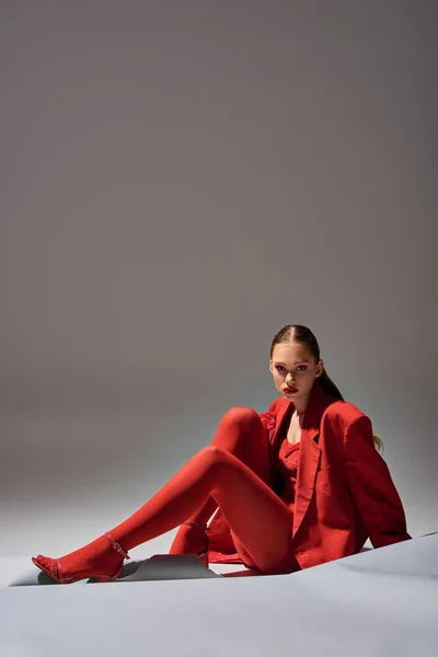 Piena lunghezza di elegante giovane donna in abito rosso con guanti e blazer posa su sfondo grigio — Foto stock