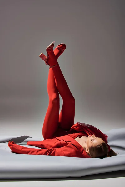 Volle Länge des stilvollen jungen Modells in rotem Outfit posiert mit erhobenen Beinen in Schuhen auf grauem Hintergrund — Stockfoto