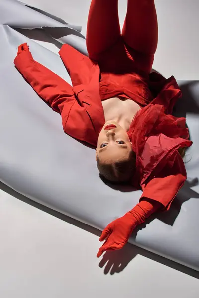 Elegante modelo joven en traje rojo con chaqueta y guantes posando con las piernas levantadas sobre fondo gris - foto de stock