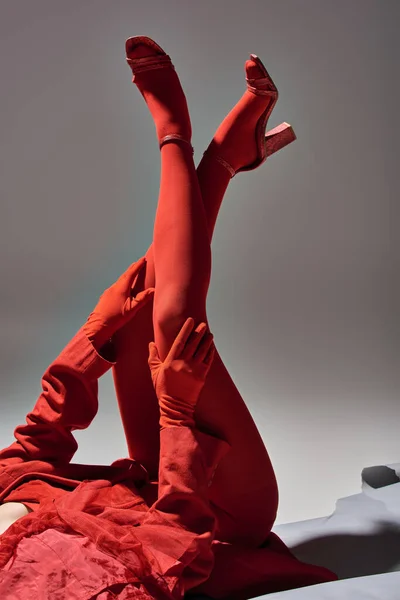 Cropped stylish model in red outfit with blazer and gloves posing with raised legs on grey backdrop — Stock Photo