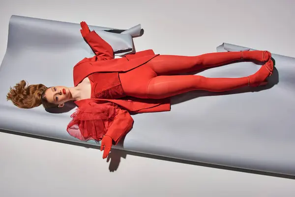 Вид сверху на симпатичную молодую модель в красном наряде, лежащую на сером фоне, женщину, действующую как дол — стоковое фото