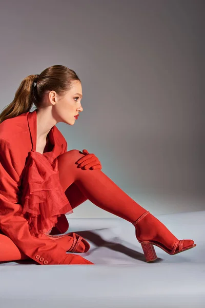Вид збоку стильної молодої моделі в червоному вбранні з колготками і високими підборами, сидячи на сірому фоні — стокове фото