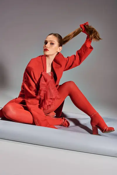 Стильная молодая модель в красном костюме с колготками и высокими каблуками тянет хвостик на сером фоне — стоковое фото
