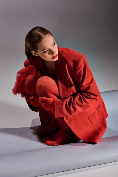 Modelo elegante em roupa vermelha com meias e saltos altos sentado e olhando para longe no fundo cinza — Fotografia de Stock