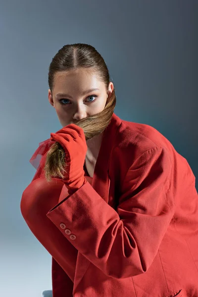 Stylish young woman in red outfit with gloves pulling ponytail on grey background, look at camera — Stock Photo
