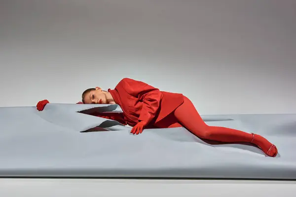 Молодая женщина в красной одежде в перчатках и колготках, лежащих рядом с разорванной бумагой на сером фоне — стоковое фото