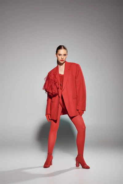 Mujer joven glamorosa en traje rojo con tacones altos y medias brillantes posando sobre fondo gris - foto de stock