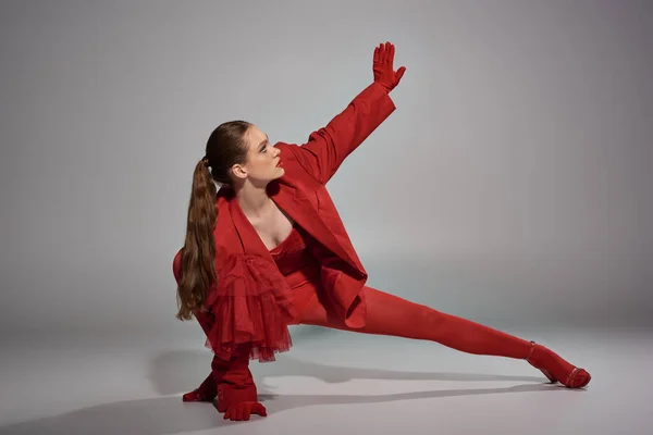 Junges Model in stylischer roter Kleidung mit Handschuhen und Strumpfhosen gestikulierend und posierend auf grauem Hintergrund — Stockfoto