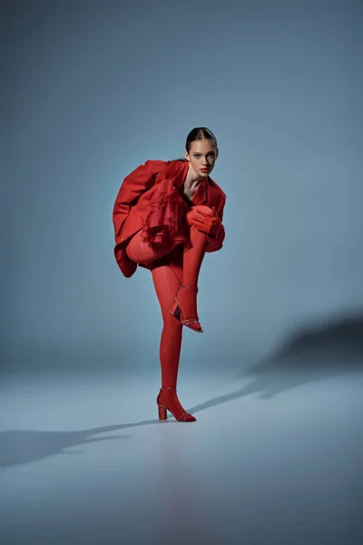 Modelo de moda en traje rojo mirando a la cámara mientras posa con la pierna levantada sobre fondo gris - foto de stock