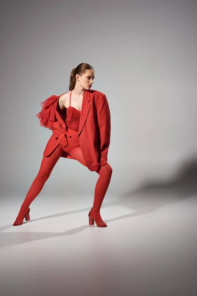 Stilvolle Frau im roten Outfit, die in die Kamera schaut, während sie auf grauem Hintergrund posiert, Modefotografie — Stockfoto