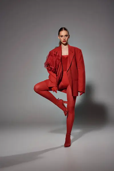 Модная женщина в красном наряде смотрит в камеру, позируя с поднятой ногой на сером фоне — стоковое фото