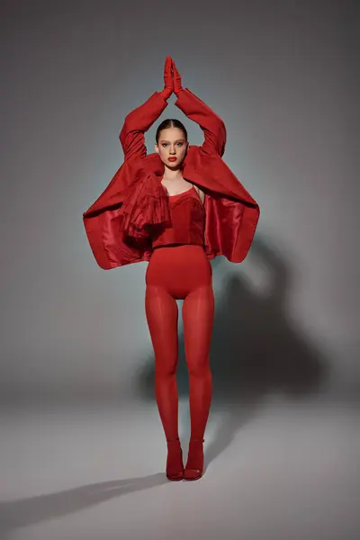 Модная женщина в красном наряде смотрит в камеру, позируя с поднятыми руками на сером фоне — стоковое фото