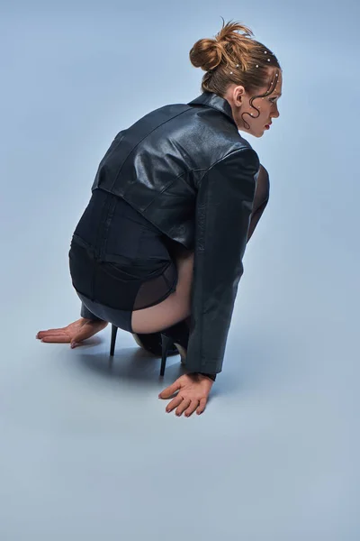 Junges trendiges Model in Lederjacke sitzt in schwarzen Strumpfhosen und High Heels vor grauem Hintergrund — Stockfoto
