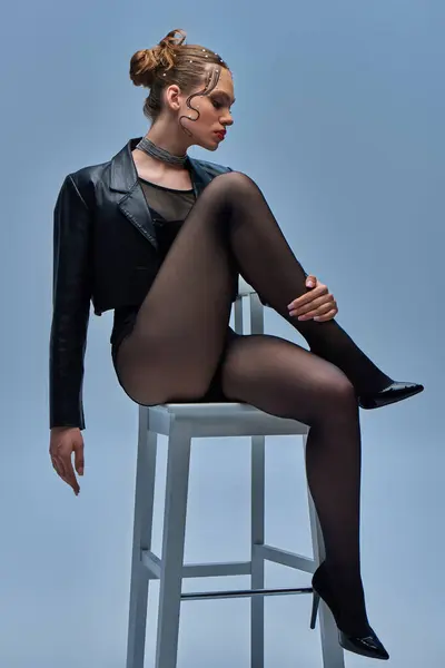 Modelo de moda joven en chaqueta de cuero negro, medias y tacones altos sentado en la silla en el fondo gris - foto de stock