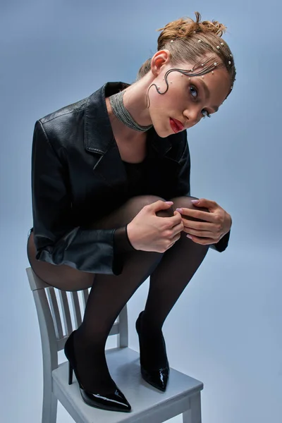 Mujer glamorosa en chaqueta de cuero, medias y tacones altos sentado en la parte superior de la silla en el fondo gris - foto de stock