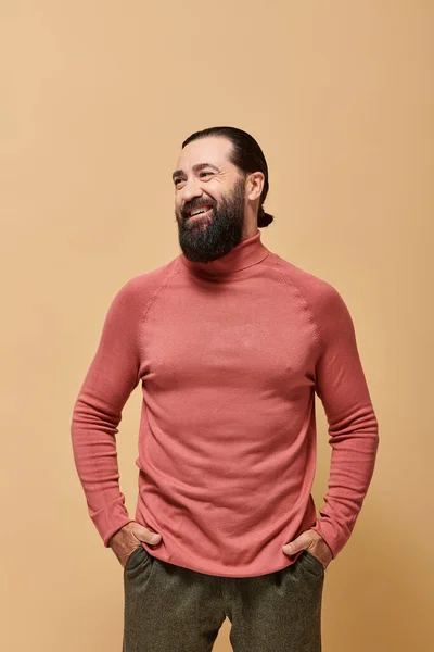 Felice uomo con la barba in maglione rosa collo alto sorridente allegramente sullo sfondo beige — Stock Photo