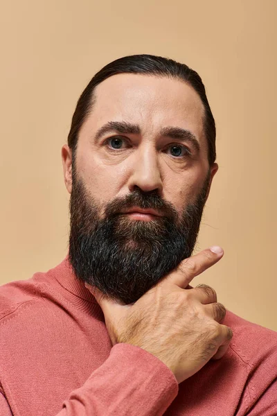 Retrato de un hombre serio y guapo con barba posando en jersey de cuello alto rosa sobre fondo beige - foto de stock