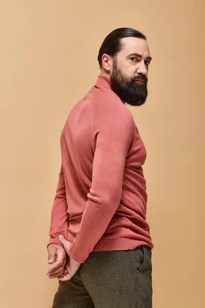 Schöner und ernster Mann mit Bart posiert in rosa Rollkragenpullover auf beigem Hintergrund, Portrait — Stockfoto