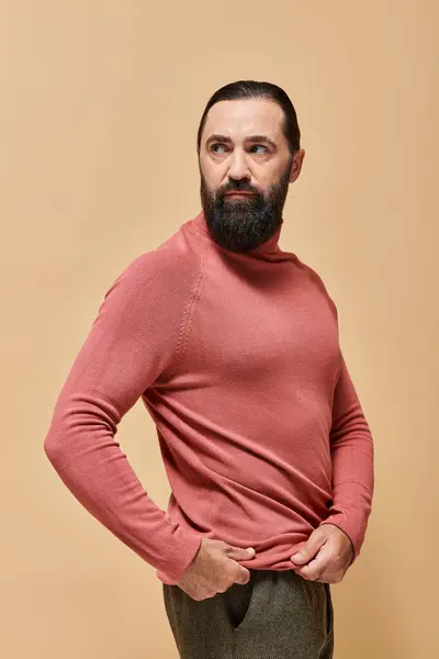 Retrato, hombre serio y guapo con barba posando en jersey de cuello alto rosa sobre fondo beige - foto de stock