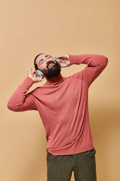 Bärtiger und glücklicher Mann im Rollkragenpullover, der Musik in drahtlosen Kopfhörern hört, beige Hintergrund — Stockfoto