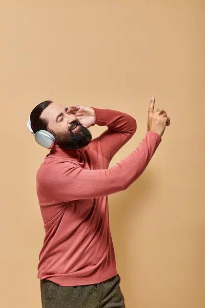 Uomo barbuto e allegro in maglione dolcevita ascoltare musica in cuffie senza fili, sfondo beige — Foto stock